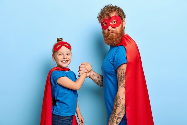 Positive female child holds hand of bearded father superhero