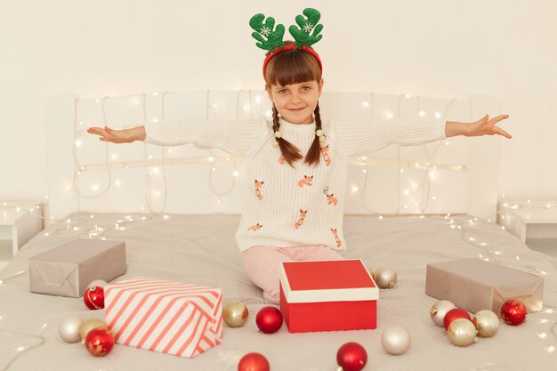 Positive female child wearing white sweater and party green deer horns sitting on decorated bed with garland and balls, posing with raised arms, looking at camera with smile.