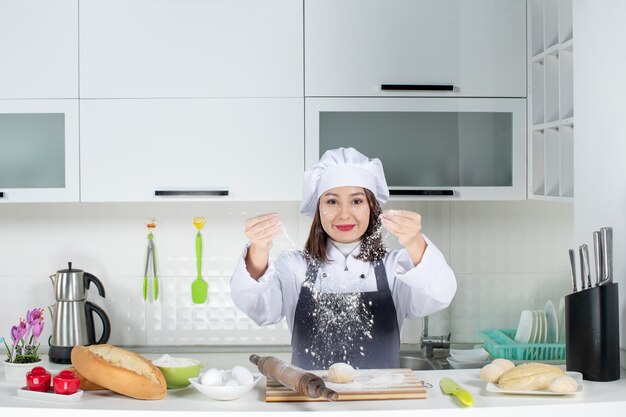 Chef commis femminile positivo in uniforme in piedi dietro il tavolo che si macchia il viso con la farina nella cucina bianca