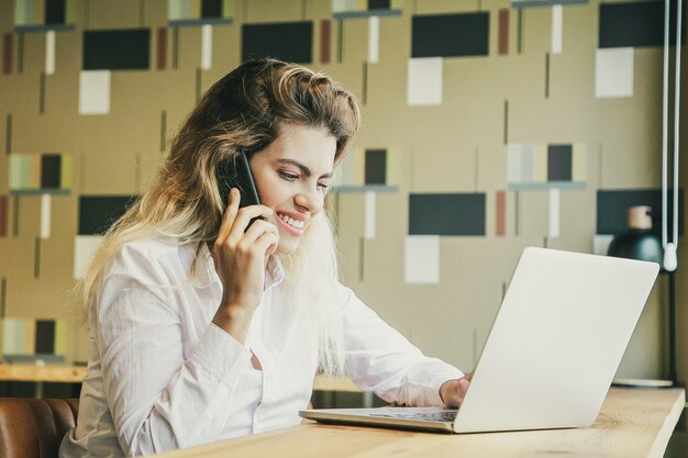 Positive female entrepreneur working at laptop and talking on cell phone in co-working space.