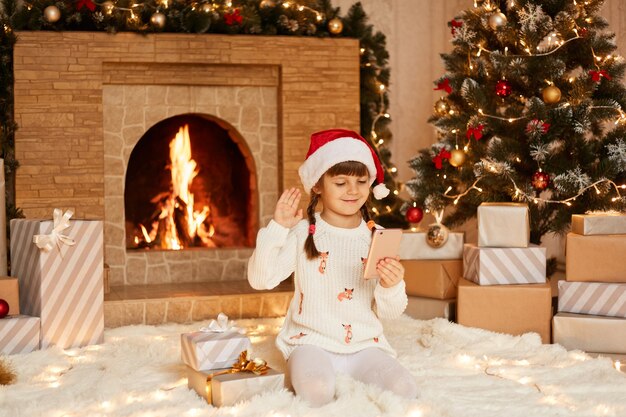 Positive female kid wearing white sweater and santa claus hat, sitting on floor near Christmas tree, present boxes and fireplace, waving hand to her friends while talking with them via video call.