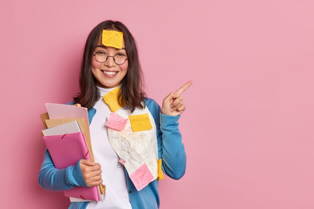 Positive female office worker with folders and stickers wears round glasses and blue jumper points away on copy space gives recommendation how to prepare successful project makes presentation