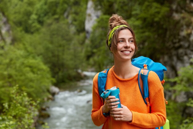 긍정적 인 여성 관광객은 나무에서 산 강 근처를 방황하고 자연 조성에 대해 포즈를 취합니다.