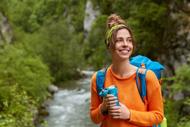 Positive female tourist wanders near mountain river in wood, poses against nature composition