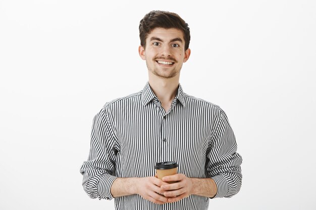 Positive friendly mature male with moustache and beard in striped shirt, holding cup of tea or coffee and smiling joyfully, meeting new people in office, talking casually and carefree over gray wall