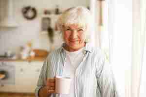 Free photo positive friendy looking senior elderly woman with gray hair and wrinkles spending day at home, drinking tea or coffee in the morning
