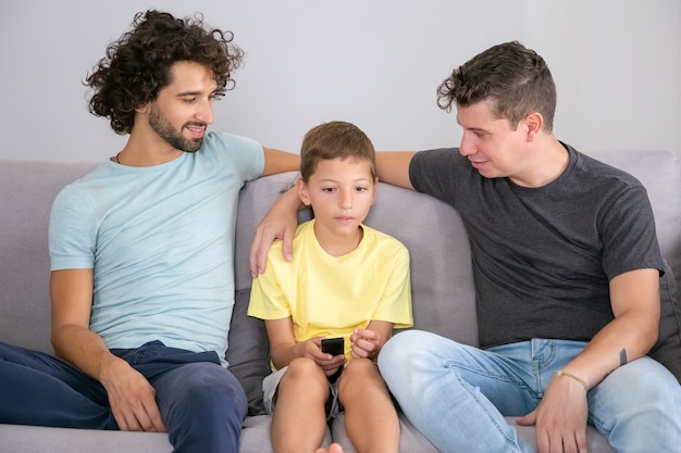 긍정적 인 게이 아빠와 아들이 집에서 함께 TV 쇼를보고 거실에서 소파에 앉아 웃고 소년을 껴안습니다. 가족 및 홈 엔터테인먼트 개념