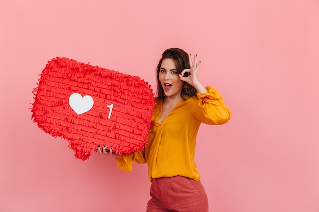 Free photo positive girl in bright blouse and trousers holds like from intagram on pink wall and shows ok sign.