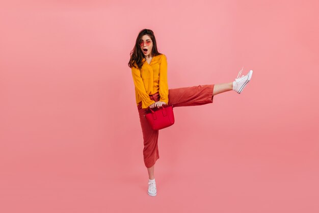 ファッショナブルな明るい服を着たポジティブな女の子は、ピンクの壁で高く跳ねます。赤いバッグと驚いたブルネットのフルレングスの肖像画。