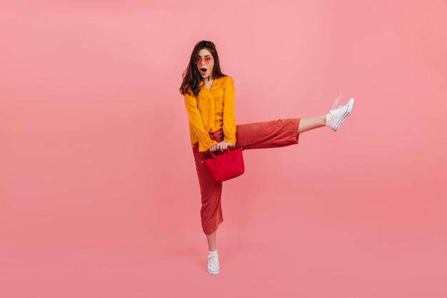 Positive girl in fashionable bright clothes bounces high on pink wall. Full-length portrait of surprised brunette with red bag.
