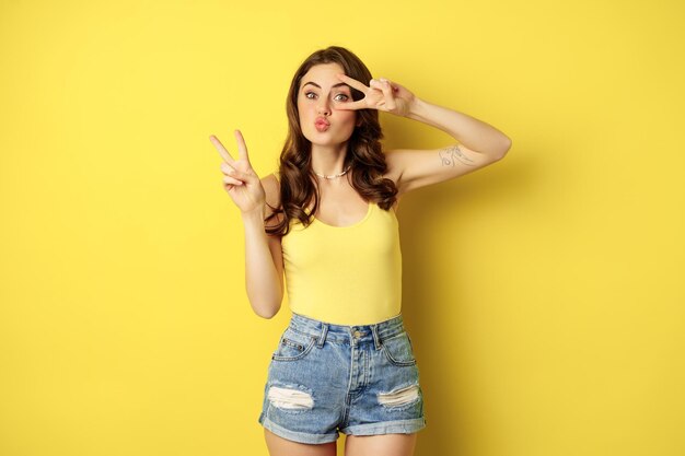 Positive girl, female model showing peace, v-sign gesture and smiling, standing in tank top and denim shorts, yellow background