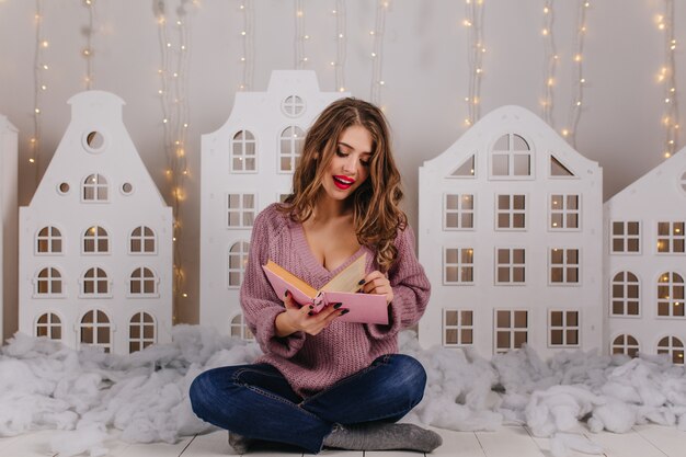 Positive girl in knitted purple sweater sits on floor and turns over page of book. Portrait of young curly-haired woman with red lipstick reading against wall of toy houses and garlands.