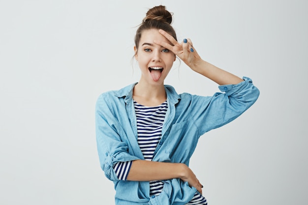 Positive girl shows her emotions. Indoor shot of attractive slender caucasian female student sticking out tongue and showing v sign over forehead, being in good mood 