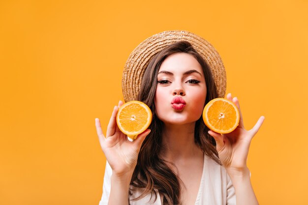 밀짚 모자에 긍정적 인 소녀는 키스를 불면, 카메라에 보이는 오렌지 반쪽을 보유하고 있습니다.