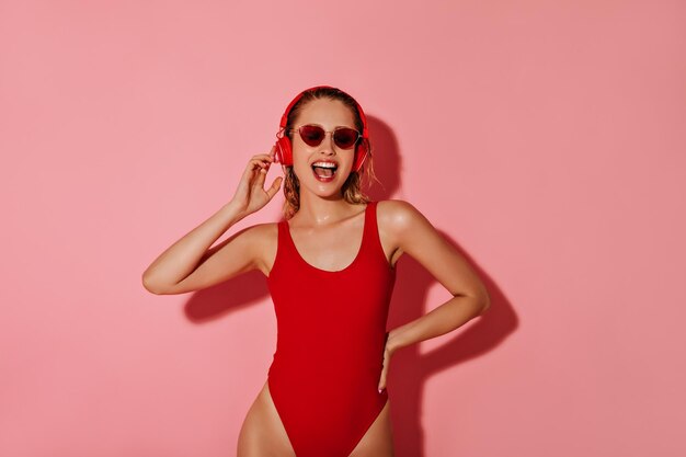Позитивная девушка с наушниками в современных красных солнцезащитных очках и ярком купальнике слушает музыку на розовом изолированном фоне