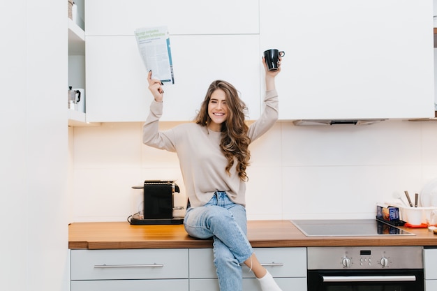 Positive girl with long dark hair holding magazine and cup of tea