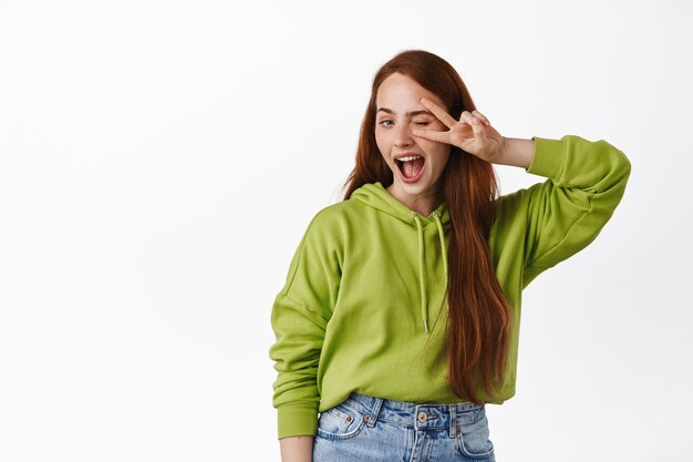 Positive girl with long natural red hair, show peace v-sign gesture near winking eye, smiling happy, posing in casual outfit on white.