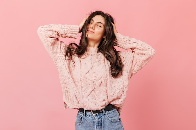 Positive girl with long wavy hair massaging her head. woman in bright outfit smiling with pleasure on pink background.