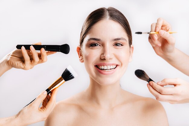 Positive girl with perfect skin is smiling, while many hands with brushes reaching for her face on isolated wall .