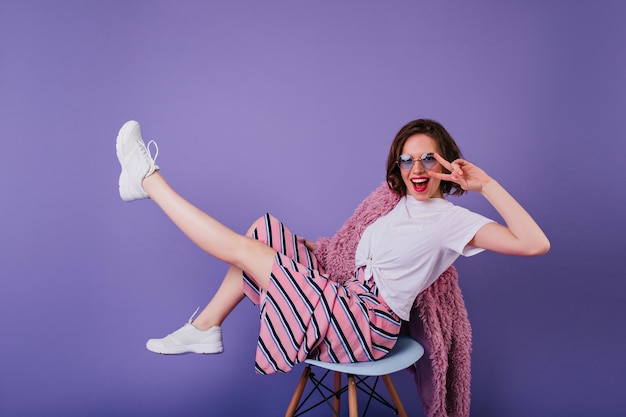 Positive girl with wavy dark hair posing on chair with peace sign Laughing woman in white shoes having fun during indoor photoshoot on purple background
