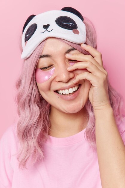 Positive glad Asian woman keeps hand on face closes eyes applies hydrogel patches under eyes removes wrinkles wears blindfold and casual t shirt isolated over pink background. Beauty concept