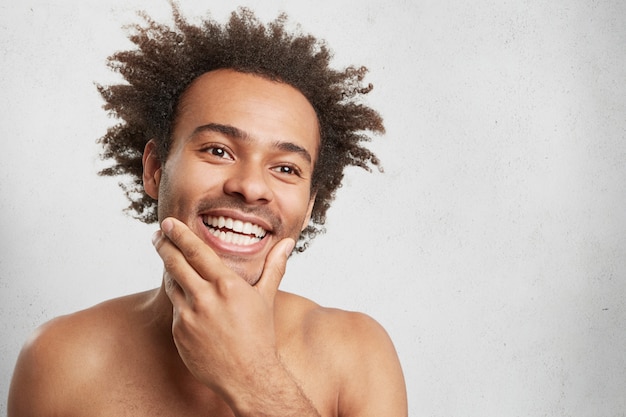 Positive glad male with dark skin, keeps hand on chin, remembers pleasant moments in life