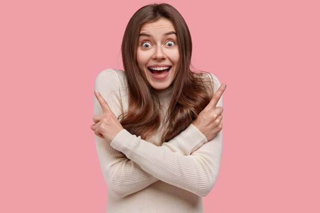 Positive glad woman keeps hands crossed over chest, points at different directions, wants two buy both things