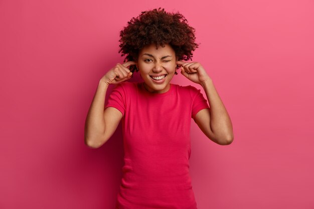 Positive glad woman plugs ears with fore fingers and winks eyes, avoids loud sound, has Afro haircut, dressed in casual wear, poses over pink wall. So noisy here. Turn off music please