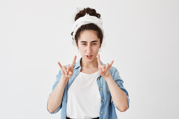 Positive gloomy young girl with dark and wavy hair in bun, wearing do-rag having fun, pouting lips, showing rock sign with both hands. Body language and gestures concept.