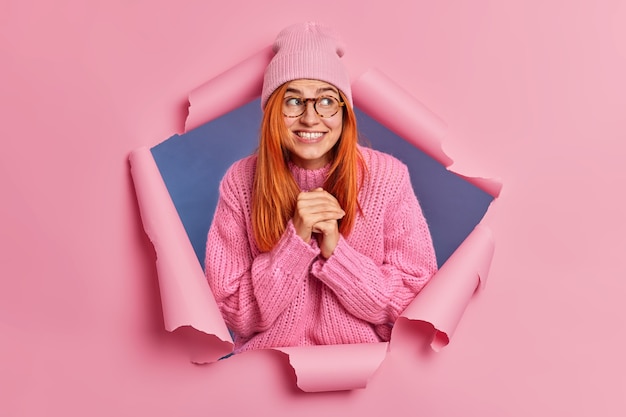 Foto gratuita la giovane donna positiva allo zenzero di bell'aspetto tiene le mani unite sorride ampiamente e distoglie lo sguardo attende che accada qualcosa di buono indossa occhiali da cappello maglione invernale.