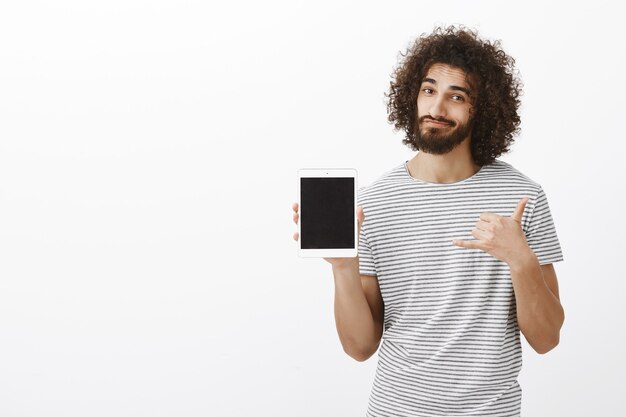 Positive good-looking male surger in striped t-shirt, showing white digital tablet and making urban gesture