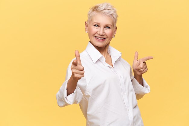 Положительная красивая европейская женщина средних лет с позированием прически пикси. Радостная дама в белой рубашке указывая указательными пальцами, рекламируя товары. Подлинные человеческие эмоции