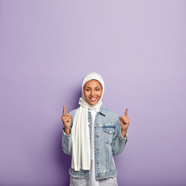 Positive good looking Muslim lady points on top, reacts on awesome promo copy space