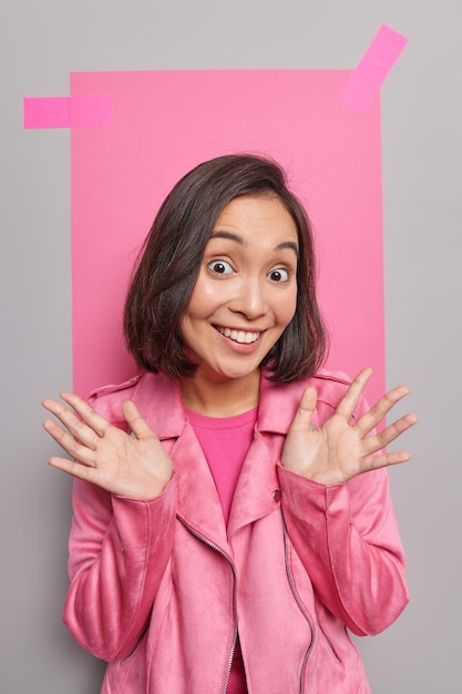 Free photo positive good looking young asian woman with cheerful smile on face spreads palms dressed in jacket meets friend stands indoor