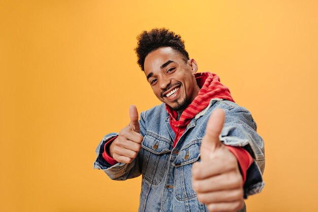 Positive guy in denim jacket laughs and shows thumbs up Snapshot of man in red hoodie smilimg and looking into camera on orange background