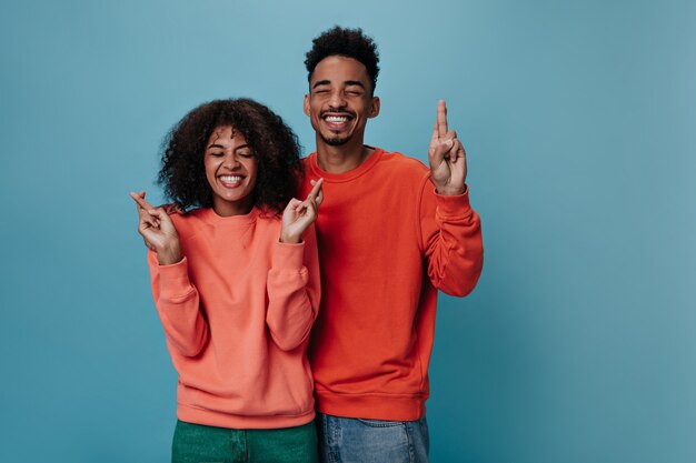 Positive guy and girl in orange sweatshirts crosses fingers on blue wall