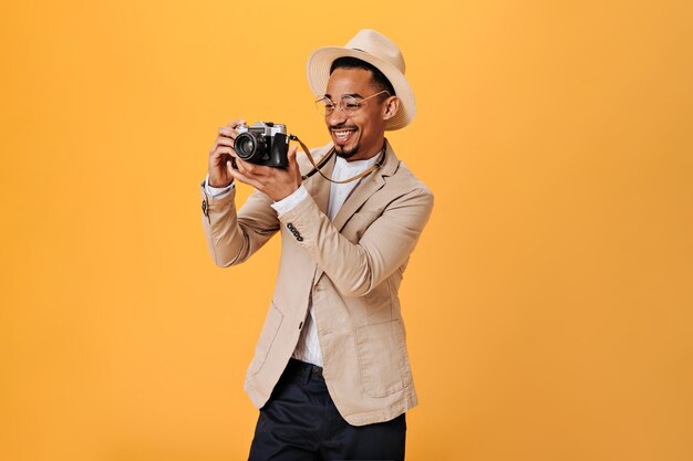 Позитивный парень в очках и шляпе держит ретро-камеру и улыбается на оранжевой стене