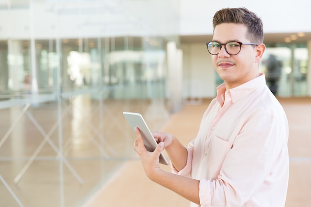 Positive guy in glasses holding tablet in office or hotel hall