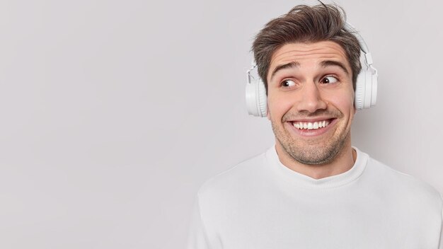 Positive guy with dark hair wears stereo headphones on ears enjoys listening music from favorite playlist smiles toothily poses against white background blank copy space for your promotional text