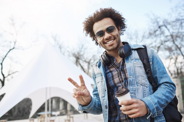 Positive handsome dark-skinned male with afro hairstyle showing peace or victory gesture while strolling across city, drinking coffee and listening music, wearing denim coat and checked shirt