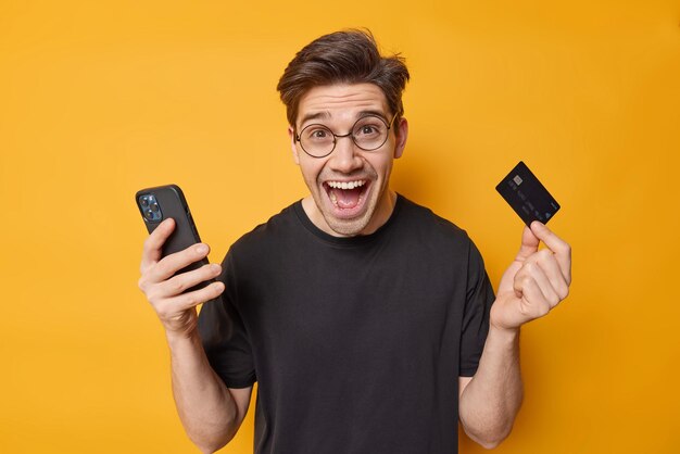 Positive handsome European man holds mobile phone and credit card makes online shopping or money transaction recommends banking dressed in casual black t shirt isolated over yellow background