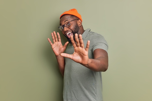 Positive handsome guy keeps palms forward front has playful mood feels upbeat dressed in casual clothes spectacles poses against dark green wall smiles happily