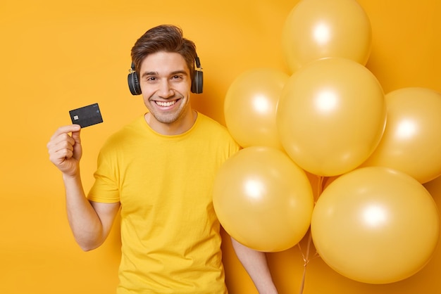 Позитивный красивый парень с темными волосами счастливо улыбается, держит банковскую карту и кучу надутых воздушных шаров, слушает музыку через стереонаушники, одетые в повседневную футболку, изолированную на желтом фоне