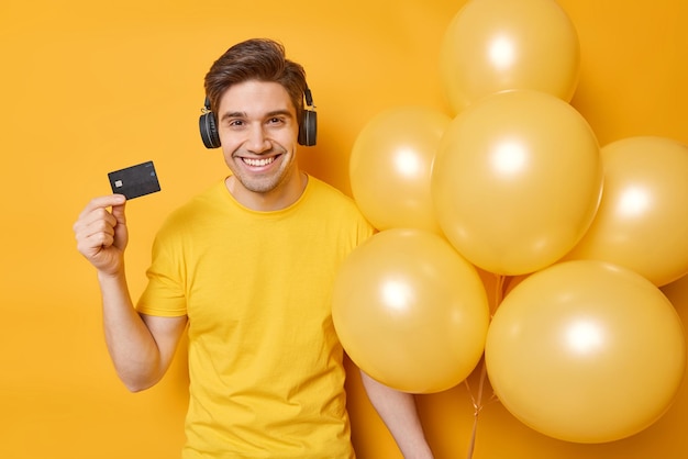 Positive handsome guy with dark hair smiles happily holds banking card and bunch of inflated balloons listens music via stereo headphones dressed in casual t shirt isolated over yellow background