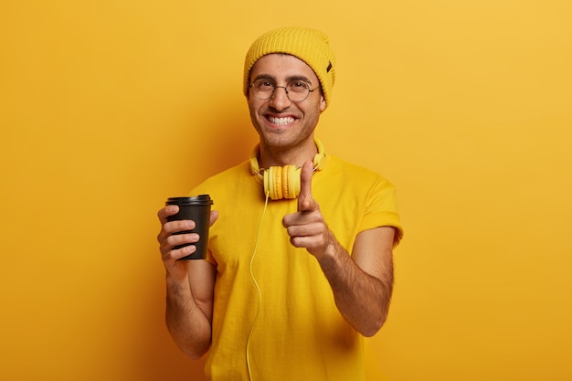 Positive handsome hipster guy points fore finger at you, makes choice, wears yellow hat and headphones
