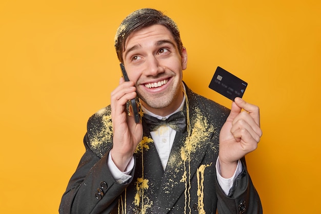 Positive handsome man makes phone call discusses latest news holds credit card dressed in festive outfit isolated over yellow background Birthday guy calls operator for getting answer on question