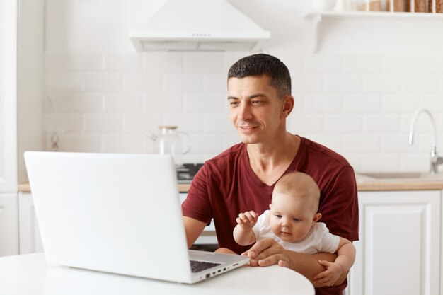 Позитивный красавец с темными волосами в бордовой повседневной футболке, смотрит в экран ноутбука, работает на ноутбуке во время присмотра за детьми, позирует на белой кухне.