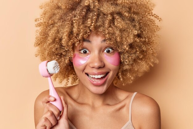 Positive happy curly haired woman with curly bushy hair uses special brush for deep cleansing applies moisturizing patches under eyes takes care of face and skin poses indoor against beige wall