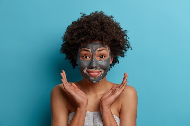 Позитивная счастливая темнокожая афроамериканка наносит маску на лицо из глины, получает косметические процедуры, ухаживает за кожей, разводит ладонями боком по лицу, стоит, завернувшись в полотенце, модели в помещении. Гигиена