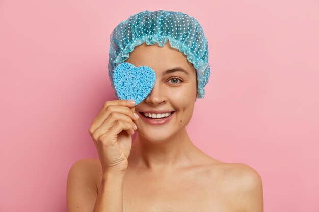 Positive happy lovely woman covers eye with heart shaped sponge, wipes face for cleanliness, wears blue protective showercap, gets beauty treatment, models against pink wall. Pampering and relax